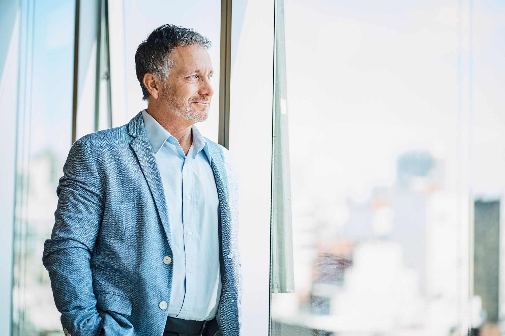 Confident male business professional looking through window. Smiling businessman is standing in coworking office. He is in businesswear.