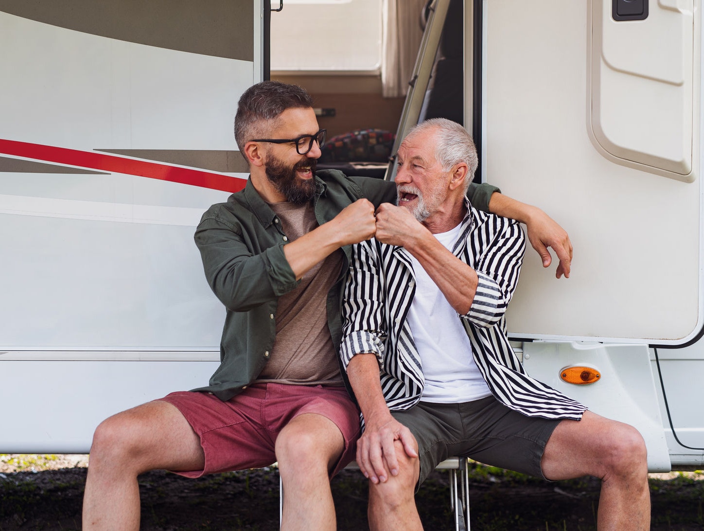 Cheerful mature man with senior father sitting by car outdoors, fun on caravan holiday trip.
