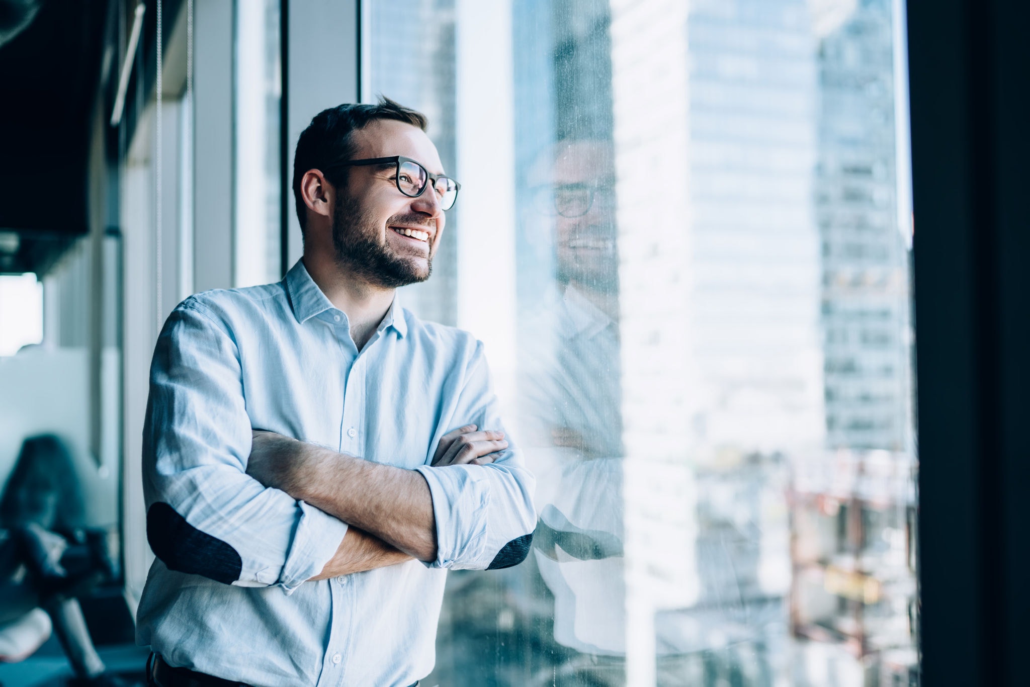Young man smiling with arms crossed, standing by an office window