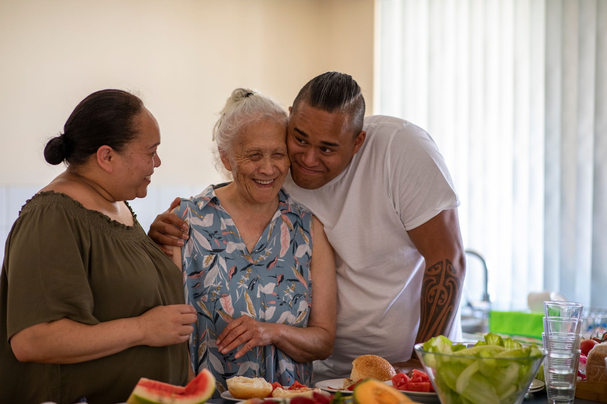 Senior Pacific Islander woman and her mature daughter and grandson cuddling each other in their kitchen at home.